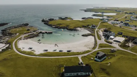 Getty Images An aerial view of a small sandy bay and harbour in Tiree. The low-lying, grassy landscape around the bay is dotted with houses and other buildings.