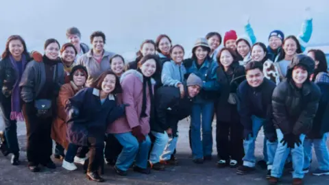 A group of Filipino nurses standing outside, what looks like a car park, they are all leaning over and smiling. One man to the right has his hands up. They are all wearing coats and warm clothing. 