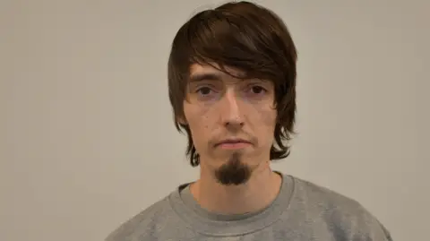 A custody photo of a man with brown hair swept across his foreheard. He is against a plain grey background, and is wearing a light grey sweatshirt. He has a short goatee beard.