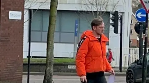 Adam Mowle wearing a bright orange jacket and dark trousers leaving Ipswich Crown Court, he has light coloured hair. 