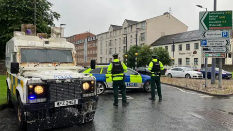 Police vehicles seal off a road in Londonderry. Two officers can be seen beside the vehicles in uniform, including high viz jackets. A number of buildings are in the background