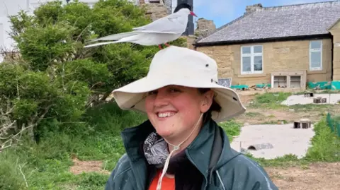 Ellen Bielinski Rebekah Goodwill on the island, in front of one of the buildings, smiling. On her head, she is wearing a sunhat. But on top of that, a Roseate Tern has actually perched on it.