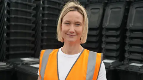Qays Najm/BBC Head and shoulder picture of Cat Sutherland looking into camera in front of black bins. She has cropped blonde hair and is wearing an orange hi-vis vest and white T-shirt.