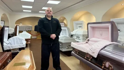 image of John Redhead standing in a funeral directors with dozens of coffins around him