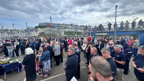 Brixham Fish Market is packed with people during the service and they have their heads bowed and are holding paper programmes. There are floral tributes on a table.