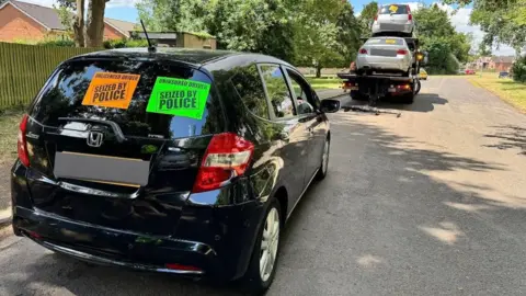 Northamptonshire Police A black car, with stickers on it, and other cars on a car transporter, on a street, with trees on it. 