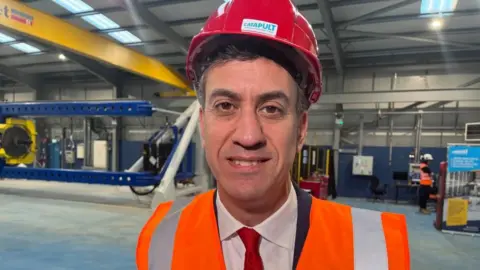 BBC Energy secretary Ed Miliband wearing a red hard hat and orange hi-vis vest, smiling at the camera. 