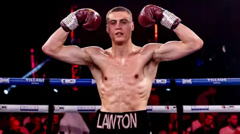 A boxer stands in a boxing ring and raises his gloved hands up to showcase his arm muscles. He has cropped dark hair, and the name Lawton is picked out in letters across the front of his shorts. People can be seen in the darkly lit background sat in seats surrounding the ring. 