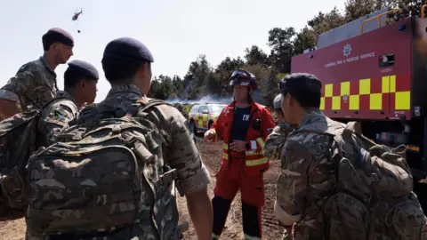 Soldiers from 10 Queen's Own Gurkha Logistic Regiment receive a briefing from a firefighter as a hleicopter with water flies overhead