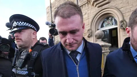 Stuart Hogg looks away from the camera outside a court. He is wearing a navy jacket and blue jumper. A policeman and journalist are beside him