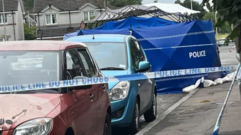 A blue police tent erected on a street. In front of it are two parked cars and some blue and white police tape.