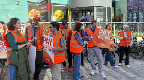 A large group of union workers and doctors wearing orange hi-vis tabards and holding plaques calling for doctors to be paid more stand outside the entrance to a hospital. They are speaking among themselves and smiling. It is sunny but they are stood in the shade.