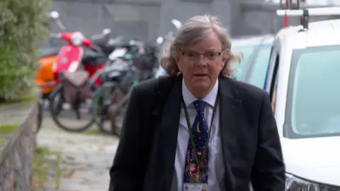 Deputy Charles Parkinson walking towards the camera. He's wearing a black suit, with a blue shirt and a Christmas themed tie. 