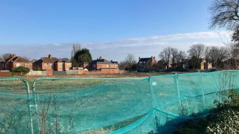 Daniel Hodgson A grassy field is bordered by a metal fence in the foreground and semi-detached houses in the background. It is is grey metal fence with a green piece of fabric pulled over it.