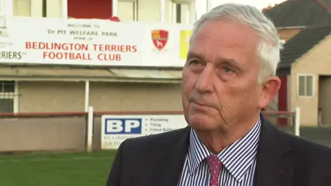 Glen Sanderson, a man in his 60s wearing a shirt, tie and jacket is standing in front of a sign for Bedlington Terriers Football Club 