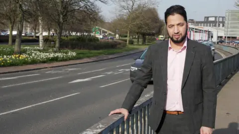 City of Wolverhampton Council A man in a suit with a pink shirt is standing next to a main road in Wolverhampton. He has short dark hair and a beard.