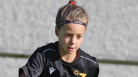 Family handout Poppy playing football. She has dark hair pulled back into a ponytail and is wearing a black football kit. She is looking down at the ground