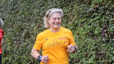 Yvonne Turner running and smiling. She is wearing a yellow top with the word 'Parkrun' on the front.