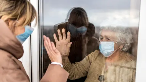 A stock photo of an older woman waving at a younger woman through the window of a care home.  They are both wearing medical face masks. The older woman has short, curly white hair and is wearing a beige jumper.  The younger woman has blonde hair tied back and is wearing a light brown hooded coat. 
