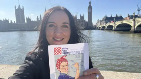 Kath Sansom Kath Sansom is standing on one side of the River Thames with Big Ben and the houses of Parliament in the background. She has long dark hair and is wearing a black blazer. She is holding a piece of paper which reds 'sling the mesh'. It has an image of a woman with her hair tied up in a red bandanna and her sleeves rolled up as is to show off her strength. It is the 'We can do it' image usually associated with women in the World War Two workforce.