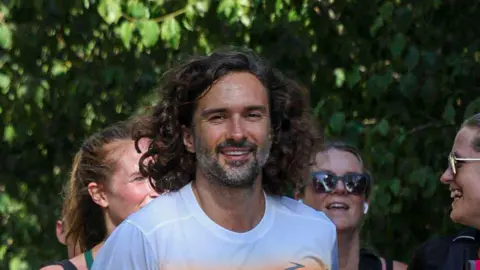 Celebrity fitness coach Joe Wicks running during a Parkrun event. There are other runners smiling and chatting around him, and he is smiling straight at the camera.