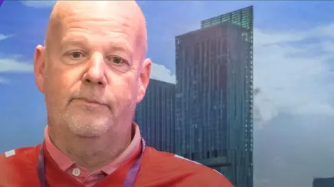 David Howarth with a short grey beard and bald head wearing an orange shirt and red jumper and a high-rise building in the background