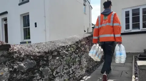 A person in hi-vis carries a number of plastic water bottles to a house.