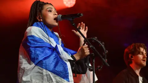 Getty Images Brooke Combe performing with a Scotland saltire flag over her shoulders. The singer is on stage singing into a microphone.