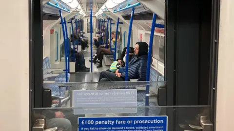 A few passengers sitting on a Tube train with their eyes closed taken through the dividing window between carriages. In the foreground there is a £100 penalty fare or prosecution sign if passengers fail to show a ticket