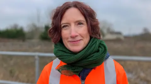 A woman with red hair stood in front of fencing. She is wearing a green scarf and an orange high-vis jacket.