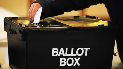 A close-up of someone reaching forward with their right hand to place a folded voting slip into the slot at the top of a black ballot box.