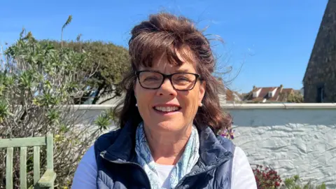 A middle aged woman with dark brown hair and black glasses smiles at the camera, wearing a blue gillet and white t shirt. in the background is a garden, trees and a house in the distance.