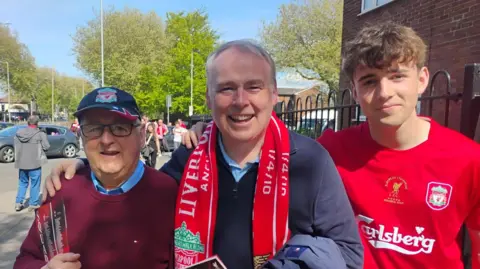 Group picture of Fred in dark baseball cap and glasses with his son in red Liverpool scarf and grandson in a Liverpool shirt