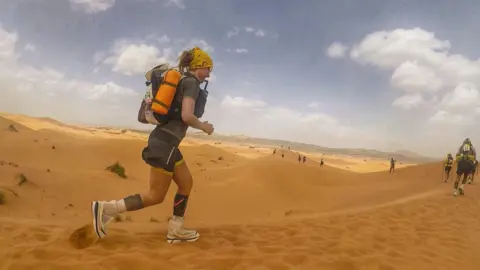 Sophie Raworth Sophie Raworth running on sand through a desert. There are other runners in front of her as they run up sandy slopes. 