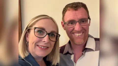 Blonde women smiling with glasses on next to a man with glasses on who's got ginger hair. They are both smiling at the camera for a selfie.