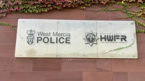 A sign on a red brick wall that reads "West Mercia Police" on the left hand side and "Hereford and Worcester Fire and Rescue" on the right. There are green and red leaves at the top of the image. 