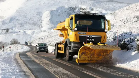 EPA A snow plough clears snow along the road with a car behind it, with snow all around on a sunny day.