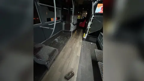 A brick is on the floor of the bus, with a broken window nearby and smashed glass all over the floor and across a number of seats.