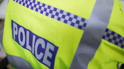 A close-up of a police high-vis jacket. The jacket is bright yellow, with the word "police" in grey, on a blue background.