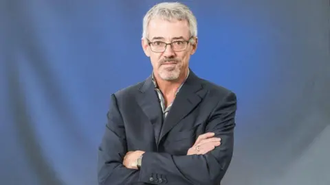 David Mearns stood against a blue backdrop with his arms crossed. He is wearing glasses and a black suit jacket, and is looking directly at the camera. 