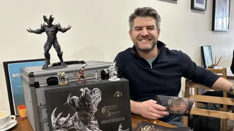A man with short dark hair and a beard in a navy blue jumper sitting behind a table with a black box, a silver container and a number of painted figures on top of them