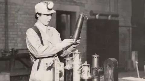 A woman stands in front of six miner lamps in what appears to be a work station. She holds one between her hands, appearing to inspect it. She is in a work uniform and wears a hat with a torch.