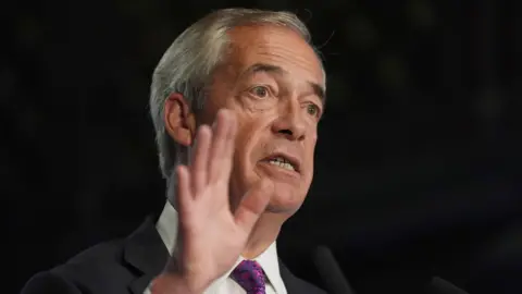 A man wearing a suit and tie with his hand in the air. He has white hair 