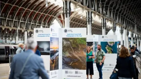 Paddington station with people walking through it. There are boards with photos the coast path with South West Photographer of the Year written on them.