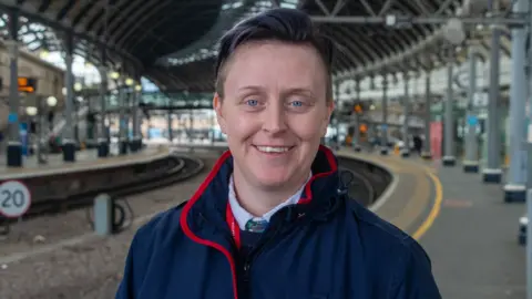 Jess Mawhinney-Coates stood on a platform at Newcastle train station, wearing a blue LNER jacket with red trim