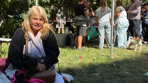 Larysa sitting on the floor holding a black cat on her handbag. People stand around under a gazebo behind her with a short terrier dog