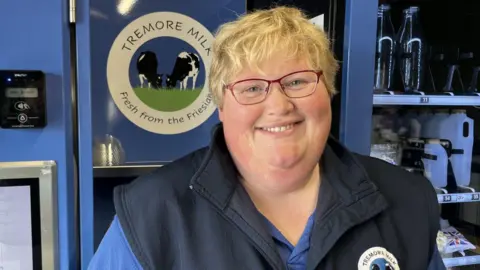 A lady with short blonde hair, blue eyes and red glasses is smiling at the camera - she is wearing a navy top and blue t-shirt underneath. She is in front of a milk vending machine.