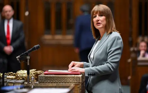Reuters Chancellor Rachel Reeves standing up in parliament. She has mid length brown hair and a fringe and is waring a grey suit jacket and white t-shirt underneath.