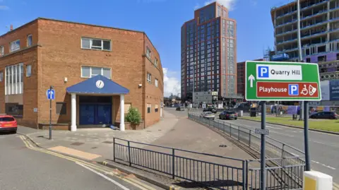 Google An urban junction in Leeds, with a three storey brick building in the foreground. The main entrance sits on the corner, marked by a blue triangular canopy on two white columns. A small clock is above the doorway. The pavement around it is wide, with dropped kerbs, paving and metal safety railings along the roadside. To the right, a large green and brown road sign stands beside the pavement and includes a blue P symbol and white text pointing towards Quarry Hill. A dual carriageway runs alongside the site, with several cars visible and traffic lights further along the road.