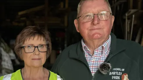 Teresa and Dave Young stood in their container. Teresa is an elderly woman with glasses and a high vis jacket. Dave is an elderly man in a green fleece and glasses.
